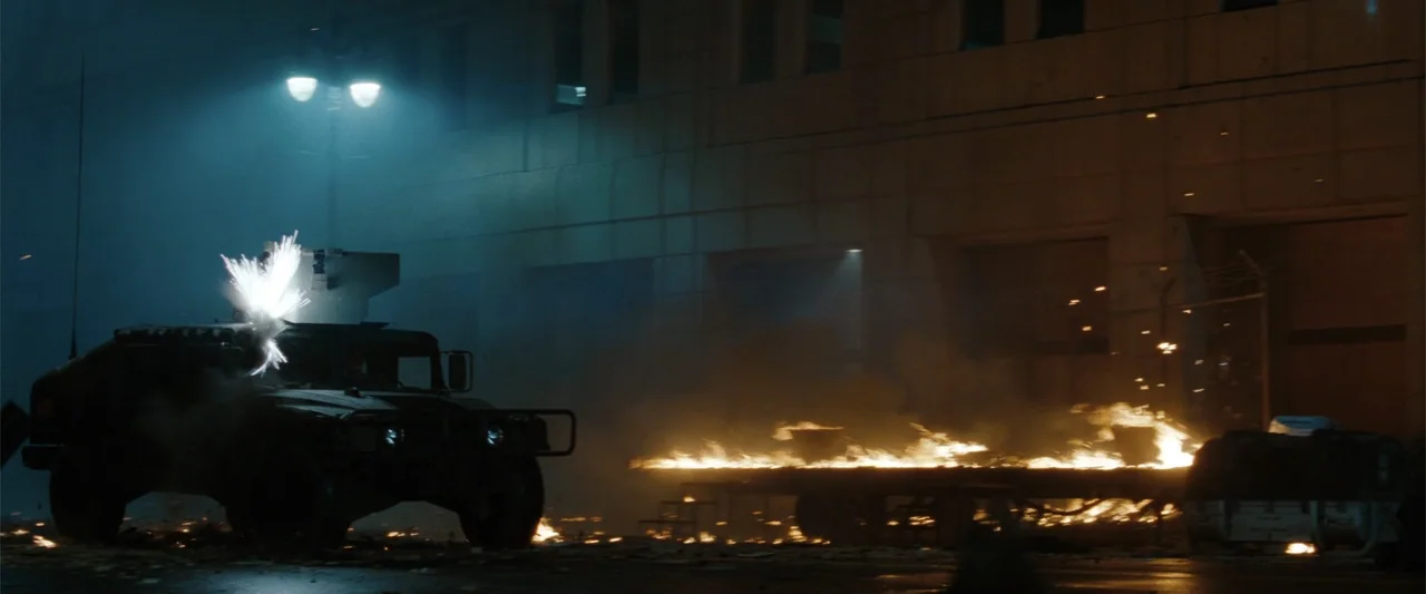 Red Dawn (2012) movie still: wide — A dark city street at night features a military humvee and a burning trailer. Sparks fly from an explosion on…