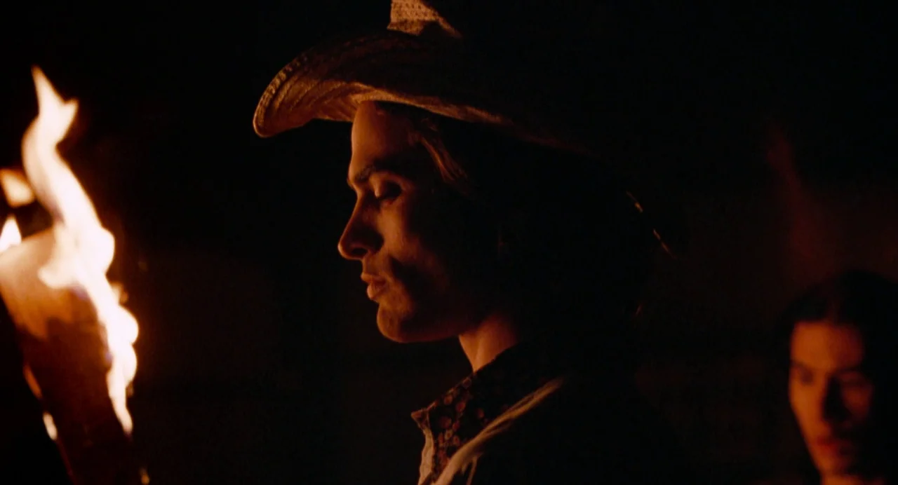 The Doom Generation (1995) movie still: medium — A dark outdoor space at night illuminated only by the light of a torch. A man wearing a cowboy hat…
