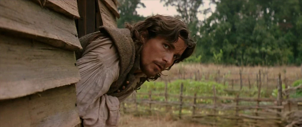 The New World (2005) movie still: medium — A rural setting with a weathered wooden structure standing next to a fenced field. A man with…