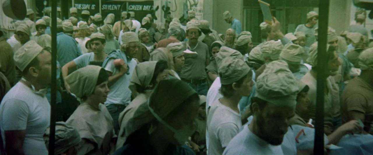 Soylent Green (1973) movie still: wide — A dense crowd of people fill a city street, many wearing simple cloth caps. The individuals are packed…