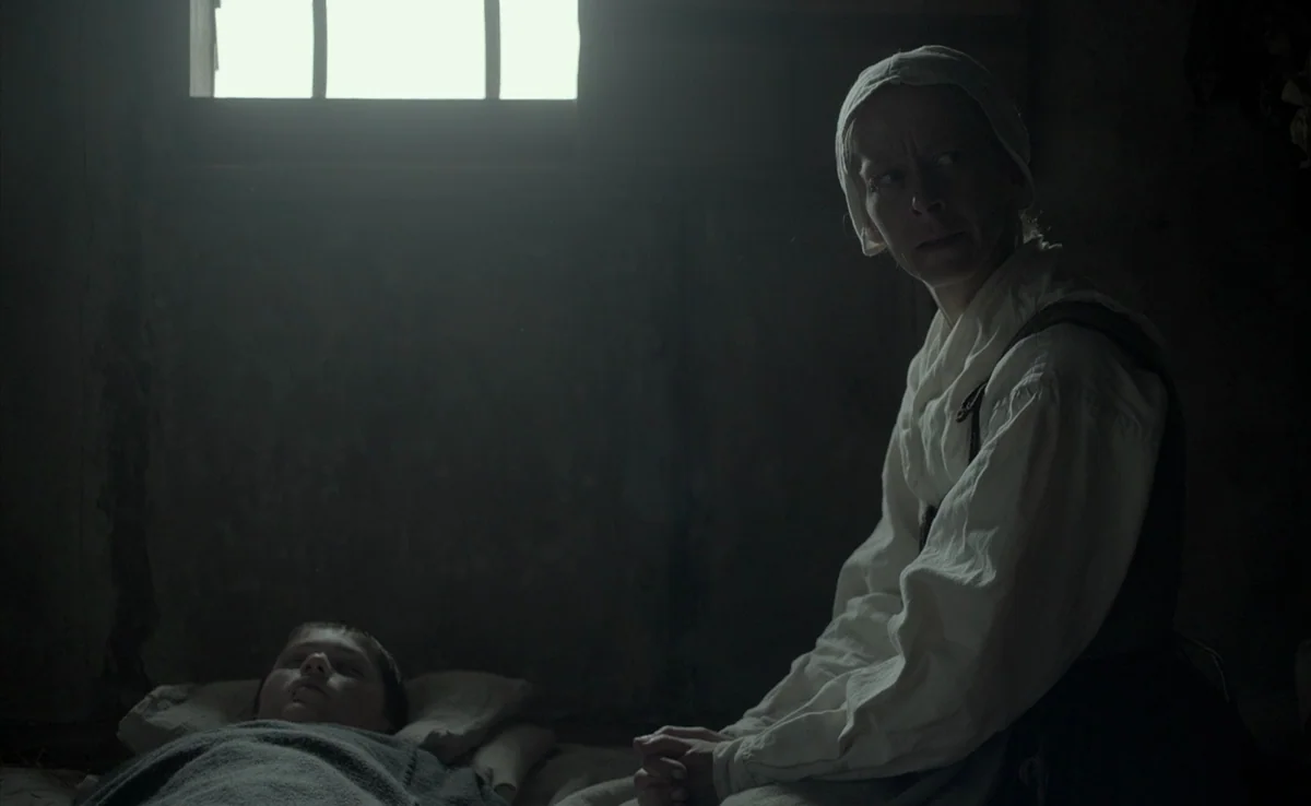 The Witch (2015) movie still: medium — A dimly lit, cramped wooden room features a small window high on the wall. A woman sits beside a young child…