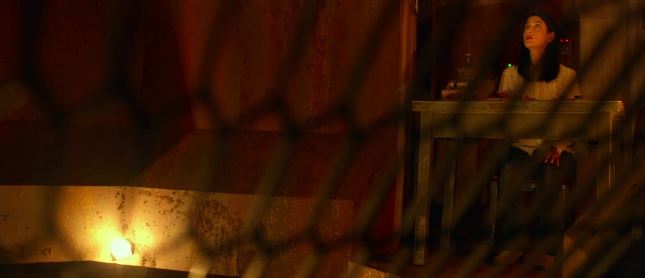 Rupture (2016) movie still: wide — A dark, rusted, enclosed space with a metal fence obstructing the foreground view. A woman sits at a small table…