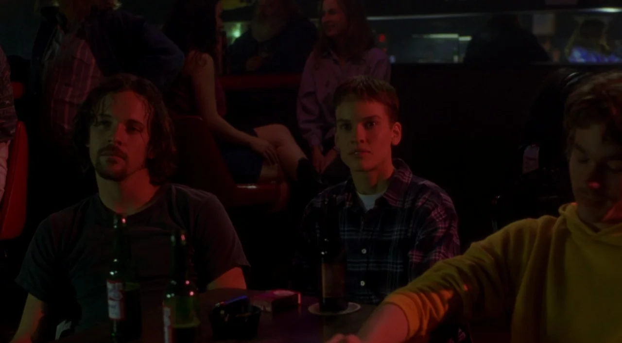 Boys Don’t Cry (1999) movie still: medium — A dark, dimly lit bar interior with people seated at tables near a counter. Three young adults sit at a…