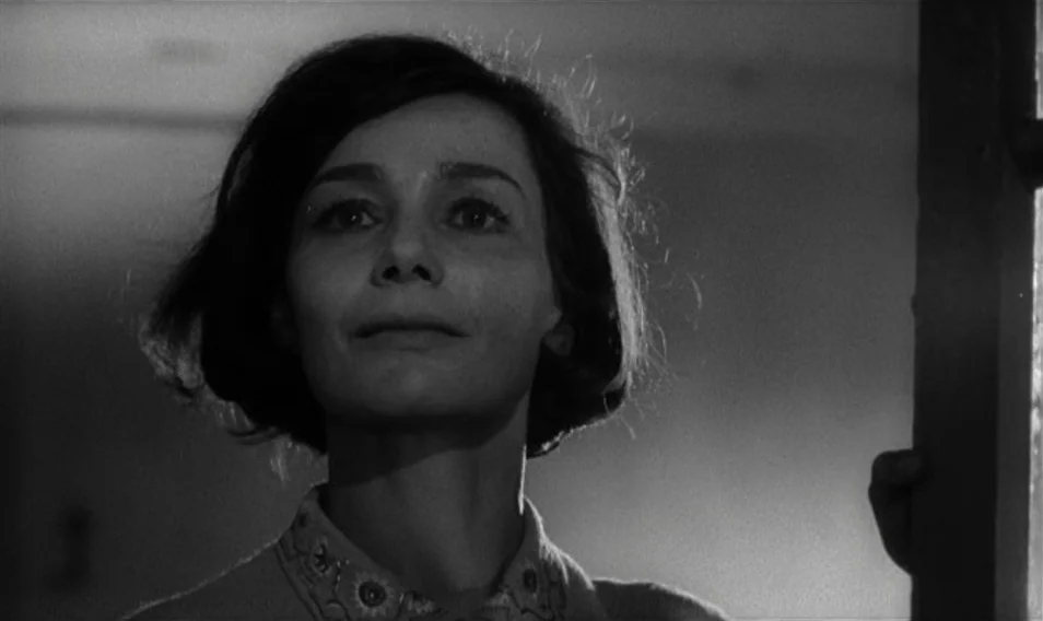 Thérèse Desqueyroux (1962) movie still: closeup — A woman stands in a dimly lit interior space with a doorframe visible on the right. She looks…