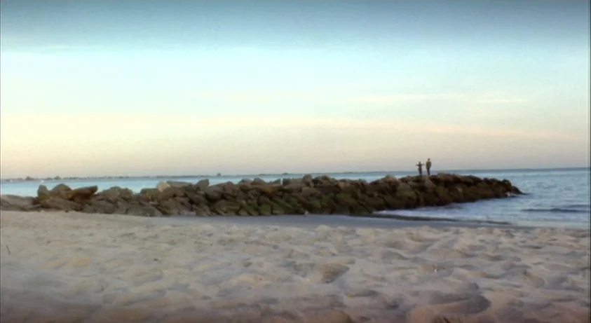 Requiem For A Dream (2000) movie still: wide — A sandy beach meets a rocky jetty extending into calm blue water at dusk. Two small figures stand…