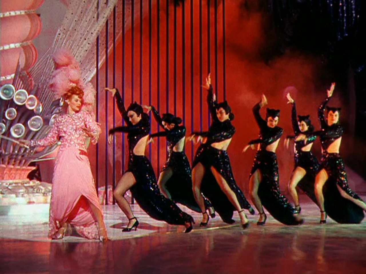 Ziegfeld Follies (1945) movie still: wide — A brightly lit theatrical stage features a red backdrop with vertical dark bars. A lead performer in…