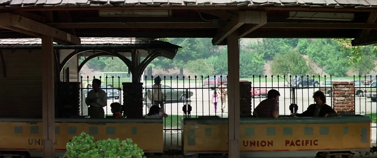 The Parallax View (1974) movie still: wide — A miniature outdoor train sits on tracks under a wooden station pavilion roof. Multiple people sit…
