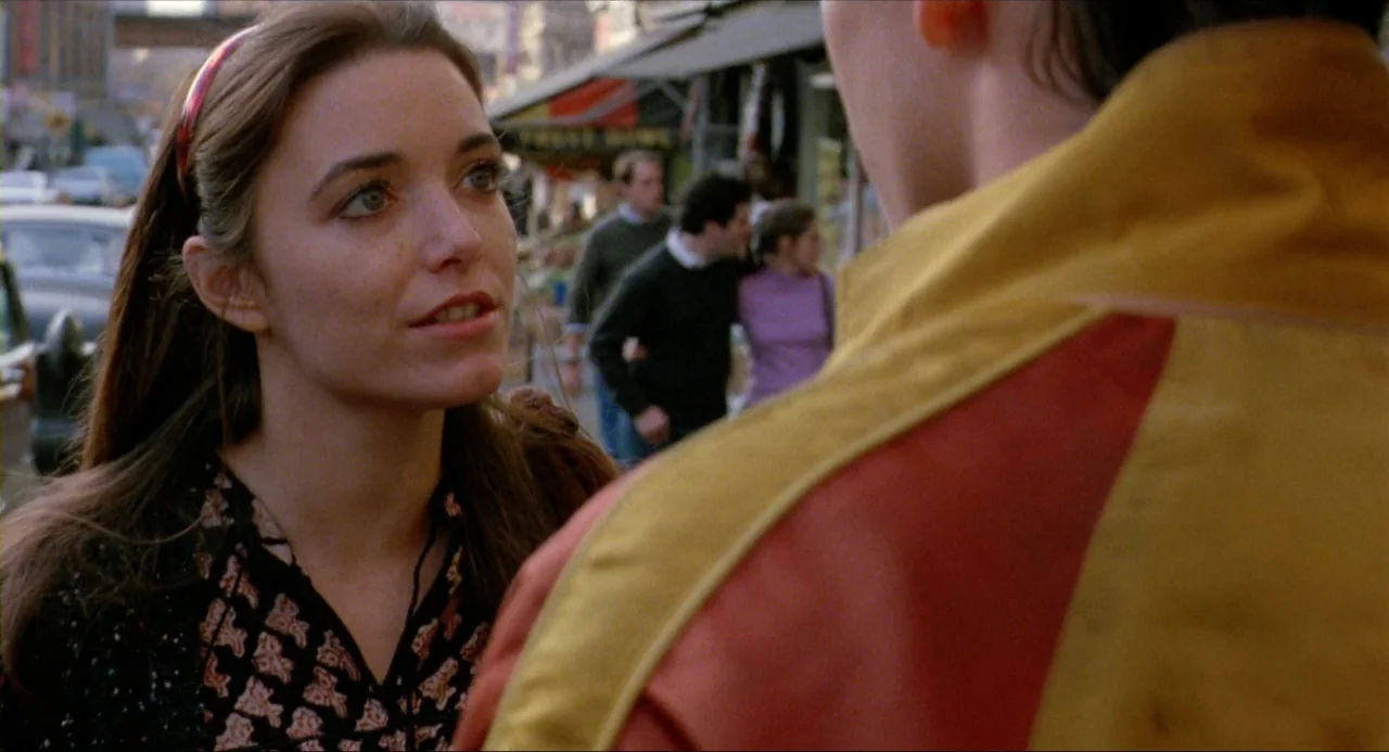 The Wanderers (1979) movie still: medium — A busy urban street with storefronts and pedestrians in the blurred background. A young woman speaks to a…