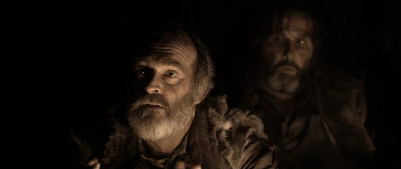 Van Diemens Land (2009) movie still: medium — Two men sit inside a dimly lit, shadowy enclosure during the night. They look upward at an unseen…
