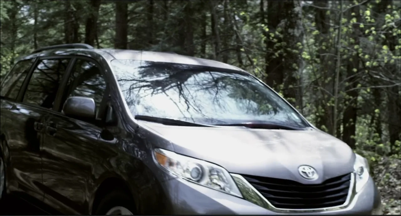 The Secret Village (2013) movie still: medium — A modern grey minivan is parked on a dirt path amidst a dense forest. The car is positioned at a…