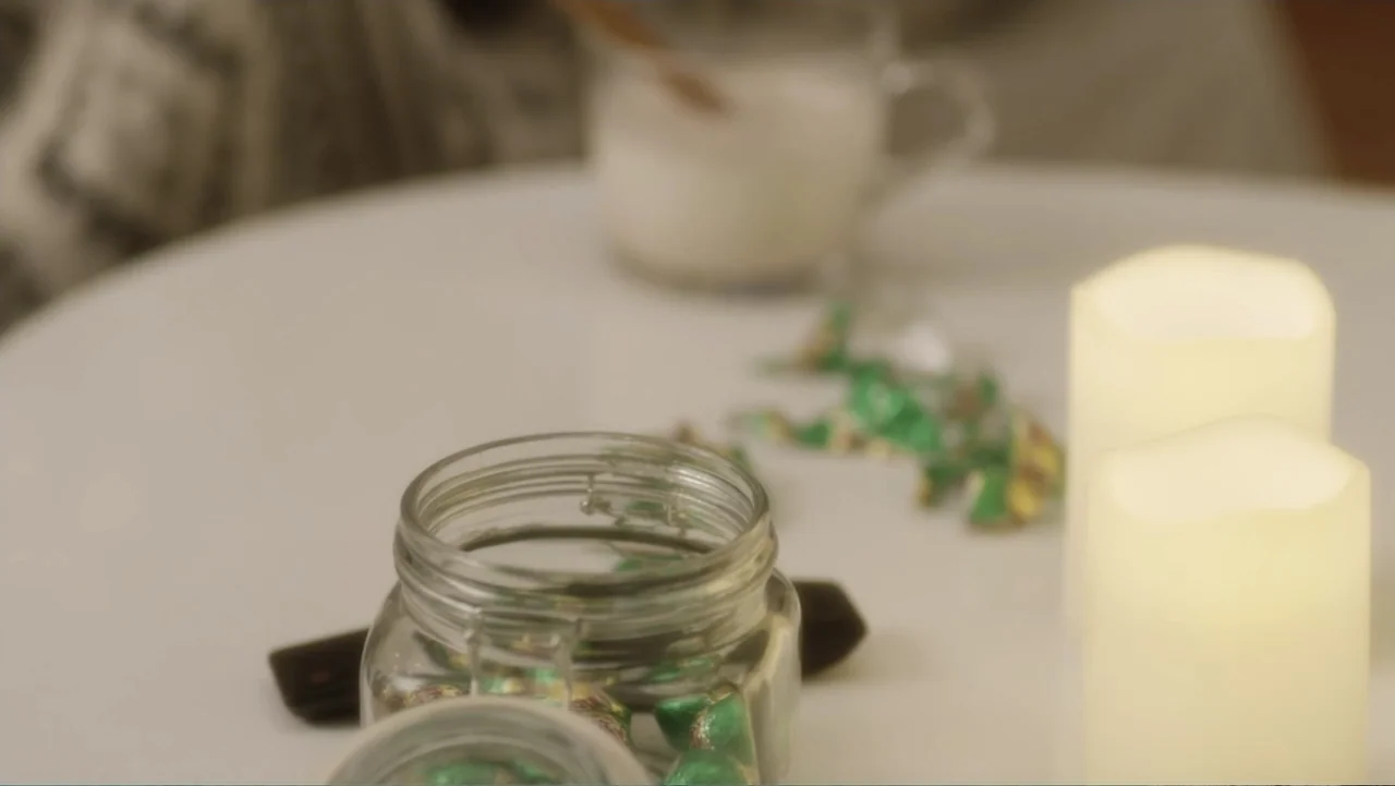 A Christmas Break (2020) movie still: closeup — A white circular table surface holds a glass jar and two battery-operated pillar candles. Several…