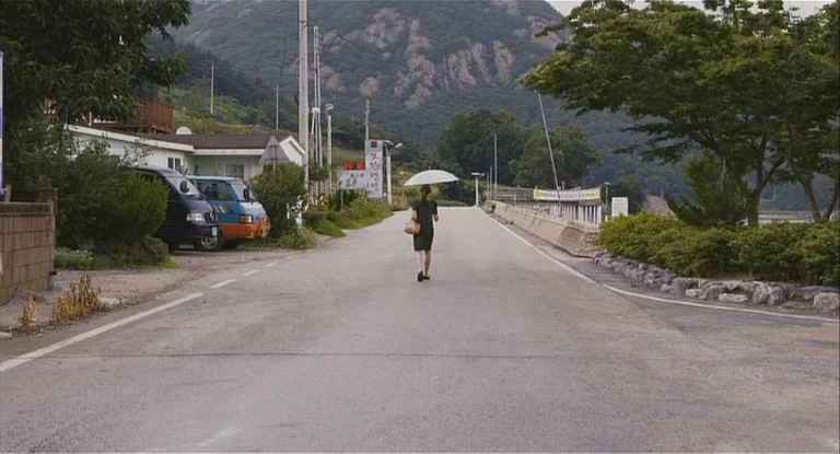 In Another Country (2012) movie still: wide — An asphalt road stretches into a hilly landscape lined with trees and sparse buildings. A woman walks…