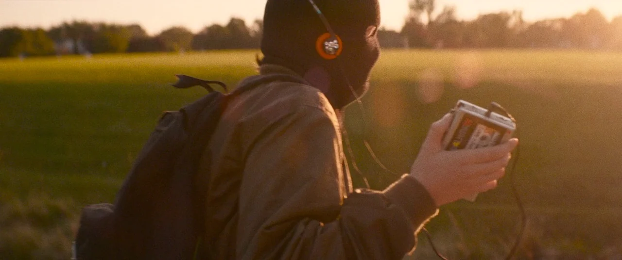 Dinner in America (2020) movie still: medium — A vast grassy field is illuminated by the warm light of sunset. A person wearing a balaclava holds a…