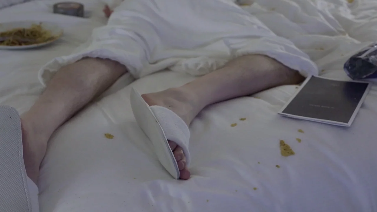 Buzzard (2014) movie still: closeup — A messy white hotel bed is cluttered with snack debris and loose items. A person in a bathrobe is lying…