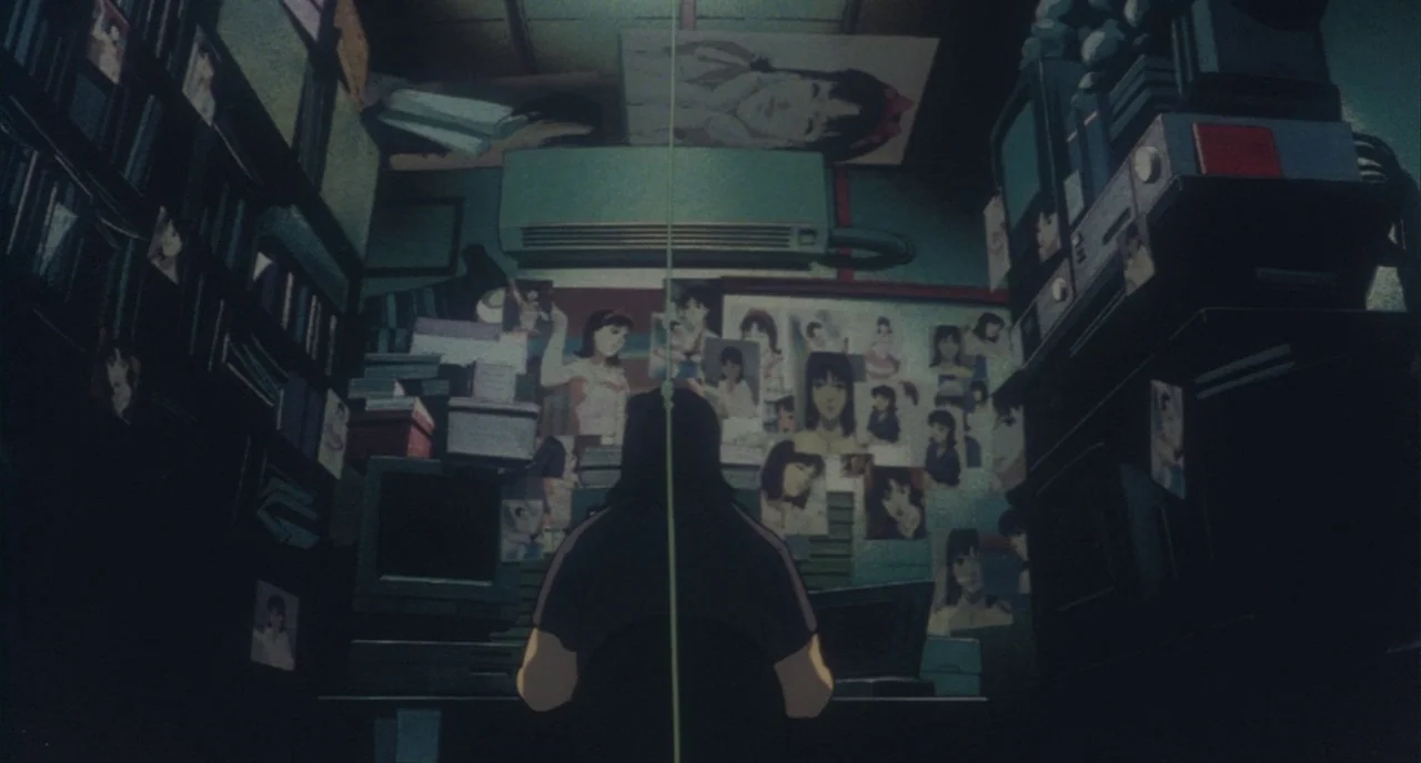 Perfect Blue (1997) movie still: medium — A cramped, dimly lit room is filled with stacks of media and technical equipment. A person sits at a desk…