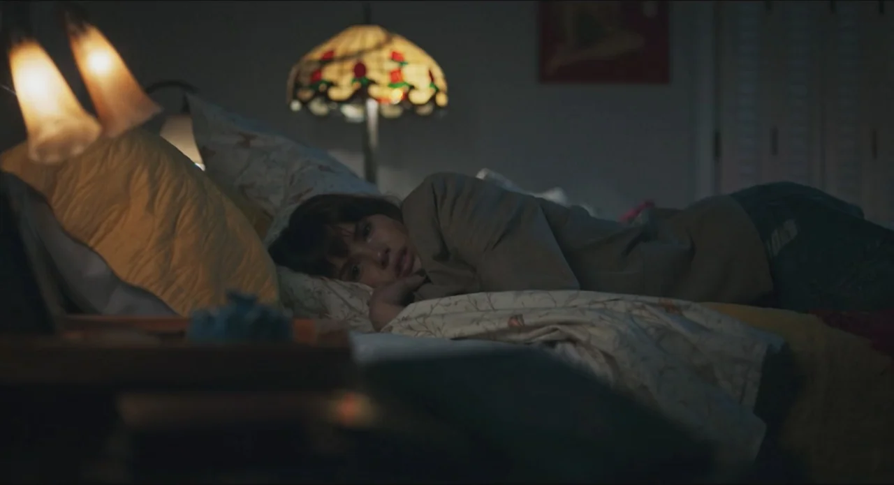 Proof of Love (2022) movie still: medium — A dimly lit bedroom features a bedside lamp with a floral glass shade. A young woman lies horizontally…