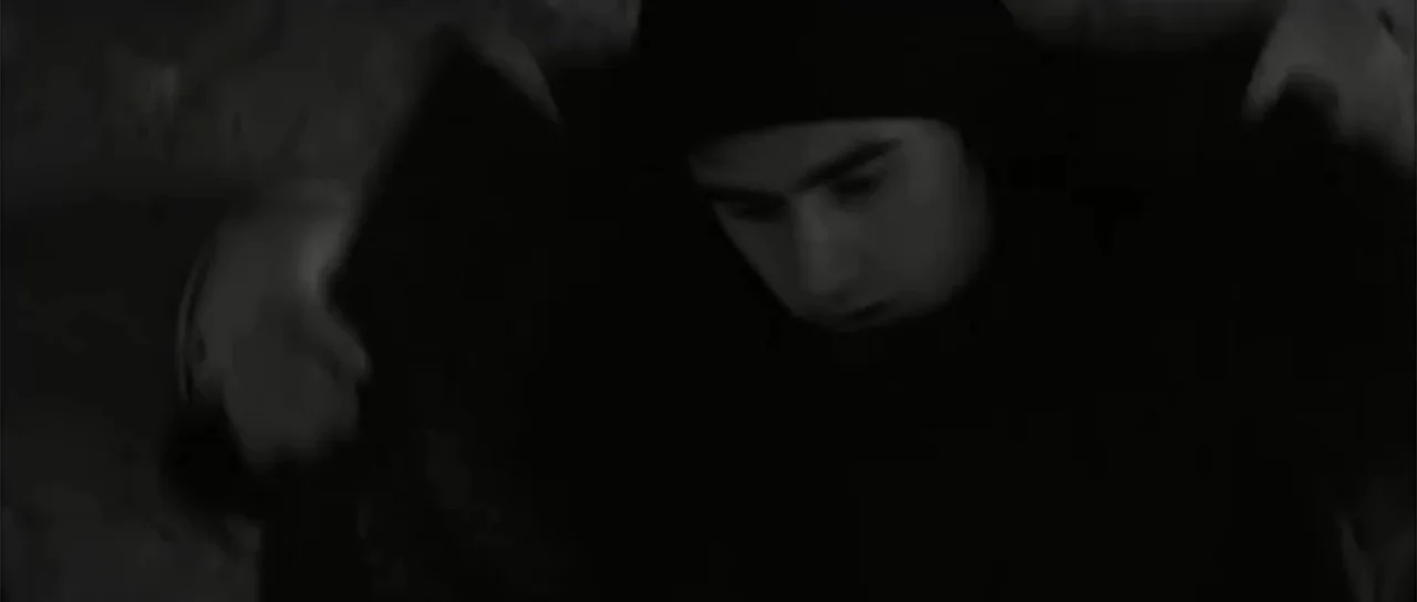 13 Tzameti (2006) movie still: closeup — A dimly lit interior space with shadows obscuring the surroundings. A young man wearing a dark hood looks…