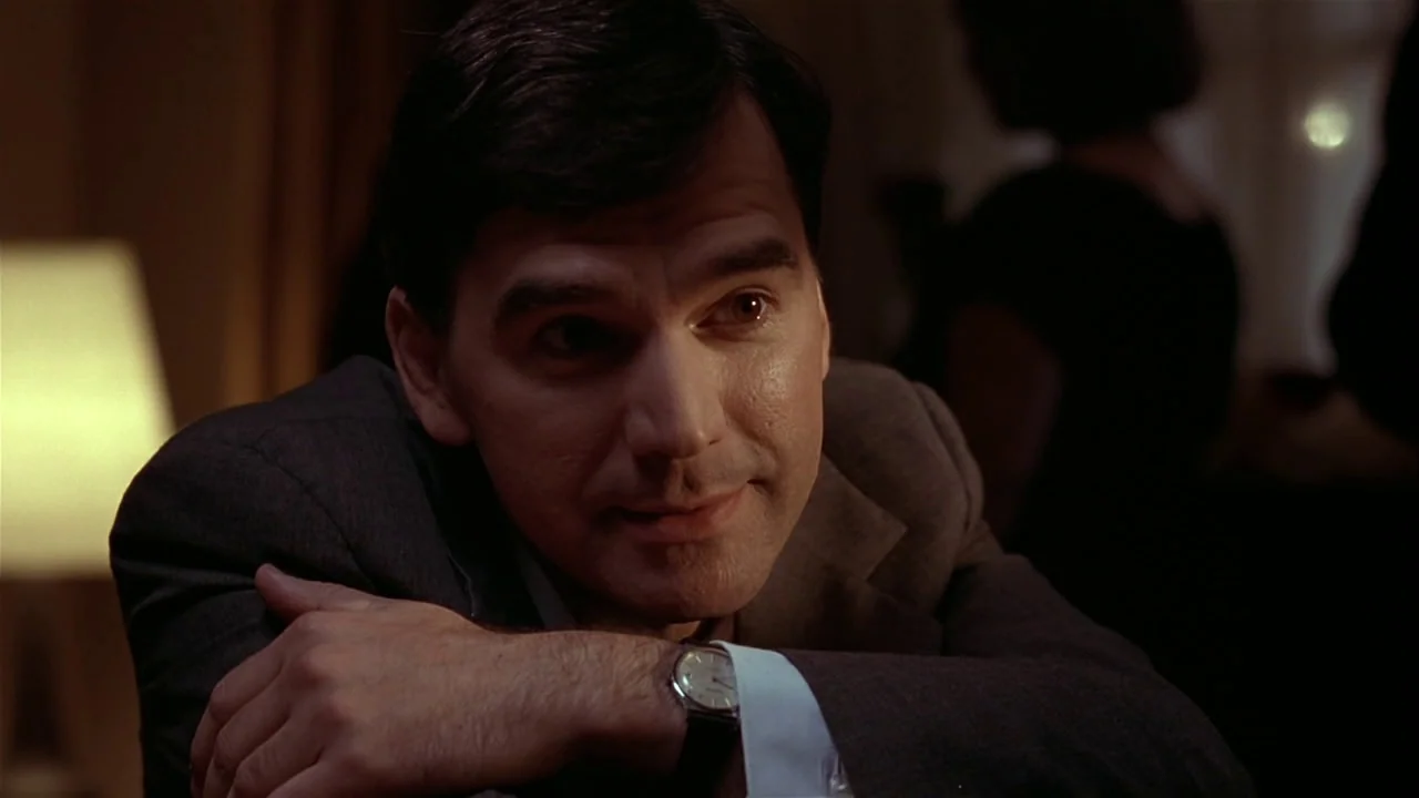 M. Butterfly (1993) movie still: medium — A dark indoor space with a lit lamp visible in the blurred background. A man in a suit looks off-camera…