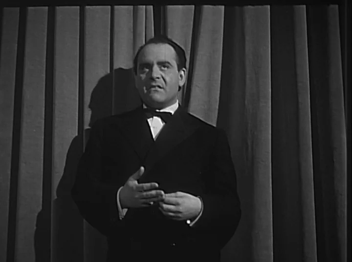 Variety Lights (1950) movie still: medium — A man stands in front of a draped curtain on a theater stage. The man is wearing formal attire and holds…