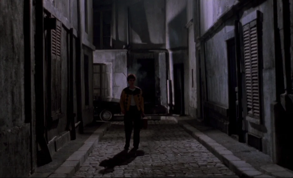 Mauvais Sang (Bad Blood) (The Night Is Young) (1986) movie still: fullbody — A narrow stone-paved alley is lined with multi-story buildings and…