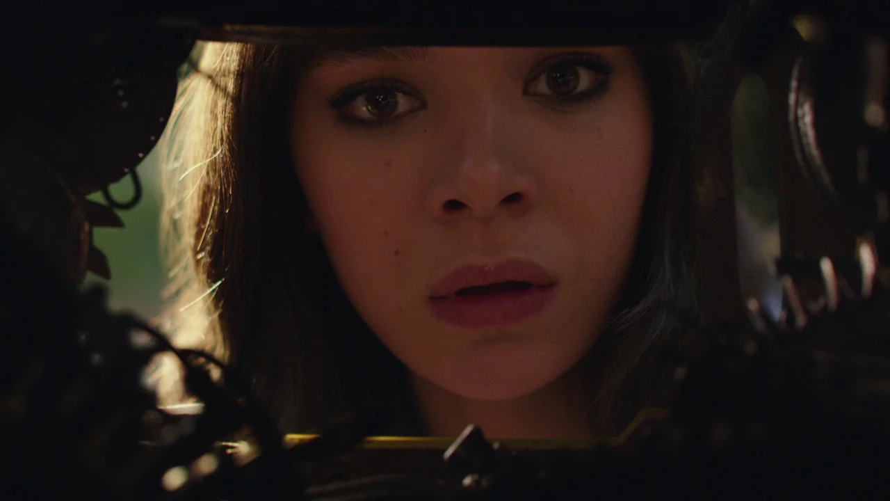 Bumblebee (2018) movie still: closeup — A woman looks through a tight dark opening framed by indistinct metal machinery components. The focused…