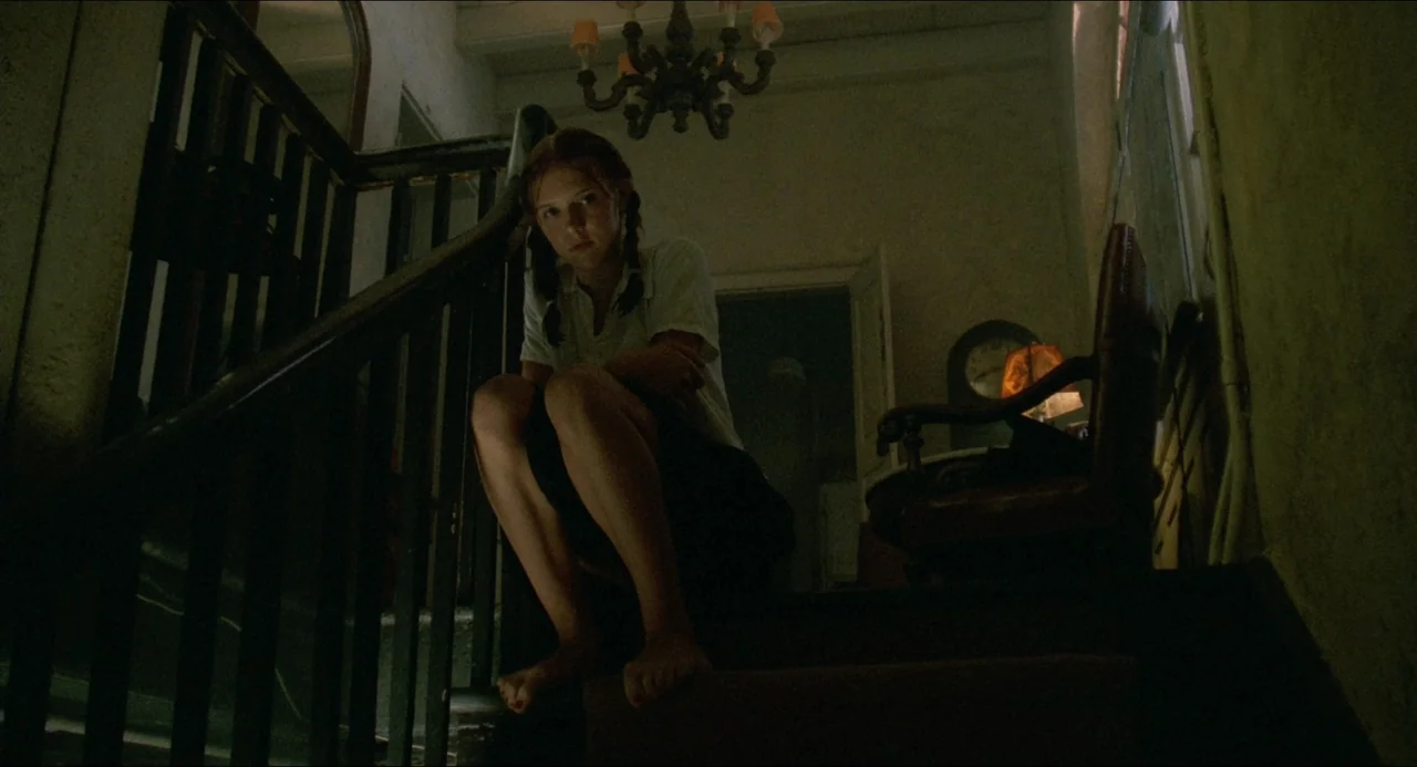 Lolita (1997) movie still: fullbody — An dimly lit staircase in an old house features wooden banisters and shadows. A young girl sits on the stairs…