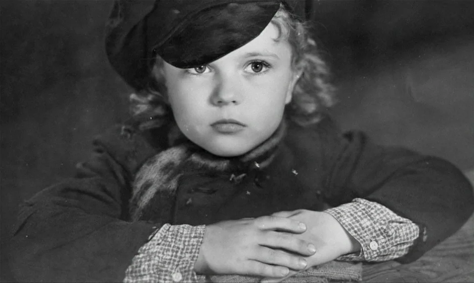 Lions Love (1969) movie still: closeup — A young child sits inside a dimly lit space with a neutral background. The child rests their chin on…