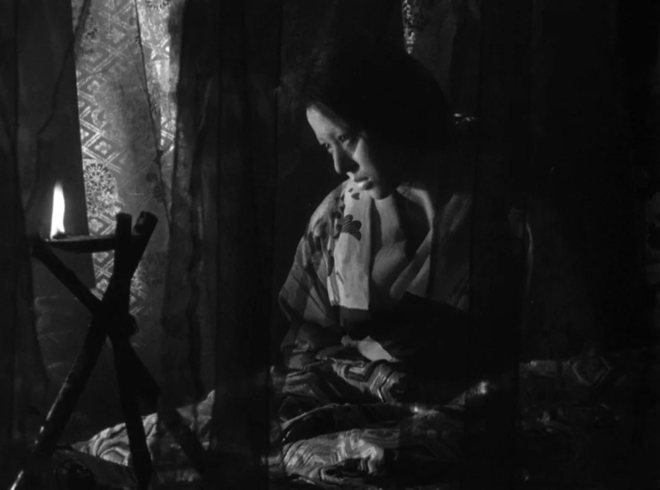 Seven Samurai (1954) movie still: medium — A dark indoor space defined by thick fabric screens and a single oil lamp. A young woman sits silently…