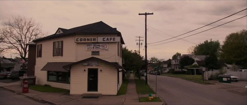 Bubble (2005) movie still: establishing — A quiet residential street corner features a two-story cream-colored building labeled Corner Cafe. Power…