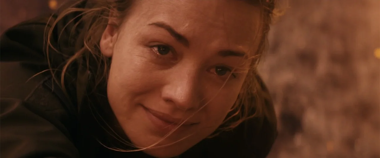 The Tomorrow War (2021) movie still: closeup — A dark outdoor setting is bathed in the warm, flickering glow of nearby fire. A young woman with…