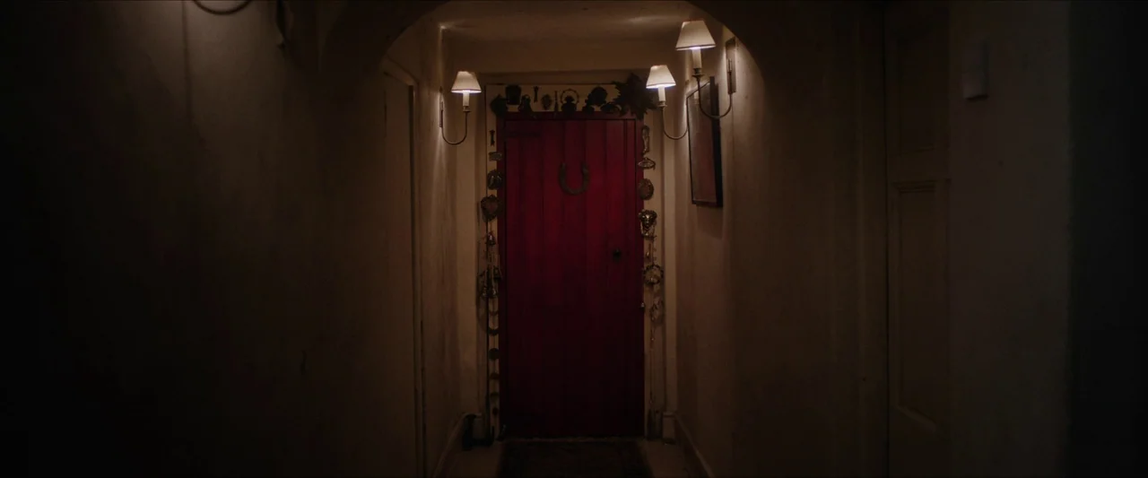 Fréwaka (2024) movie still: wide — A dark, narrow residential hallway leads to a prominent red wooden door at the end. The door frame is decorated…