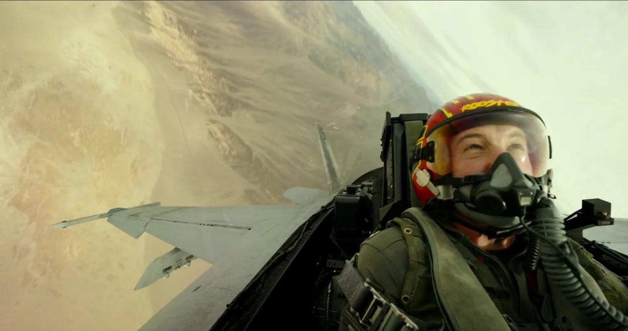 Top Gun: Maverick (2022) movie still: medium — A military jet pilot sits in the cockpit of a fighter plane. The aircraft banks steeply over a rugged…