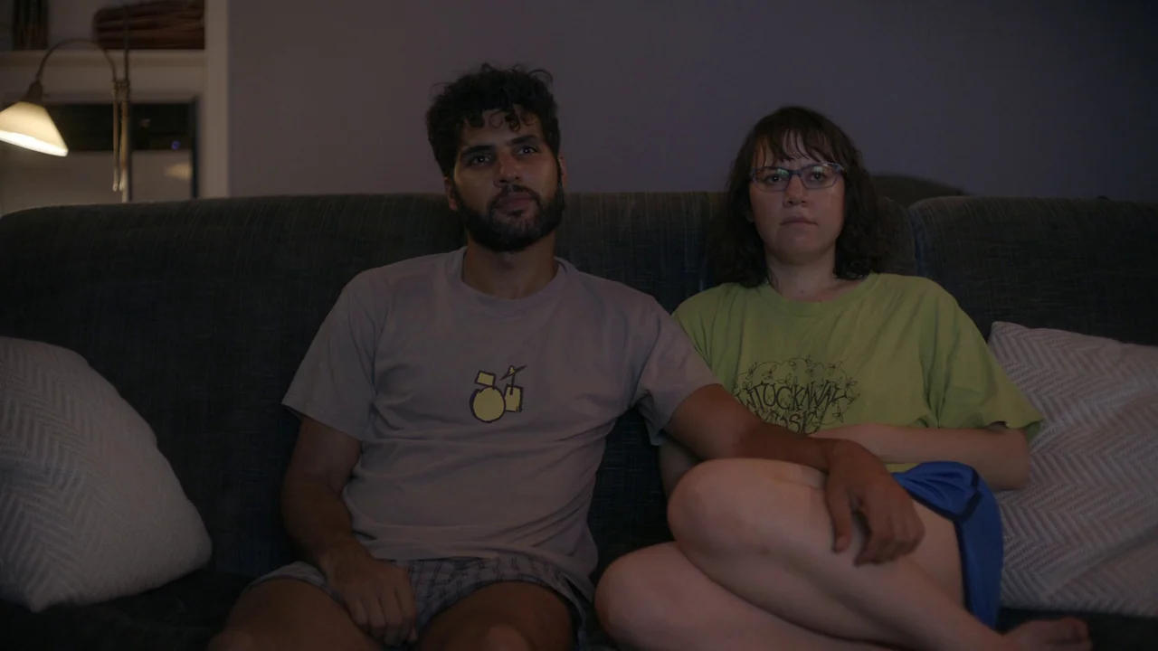 The Feeling That the Time for Doing Something Has Passed (2023) movie still: medium — A couple sits side-by-side on a dark grey fabric couch at…