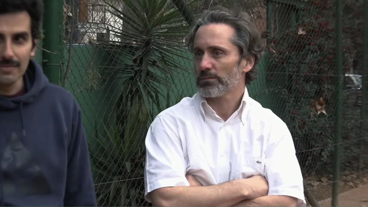 Smog in Your Heart (2022) movie still: medium — Two men stand in front of a green chain link fence with overgrown foliage. The men face toward the…