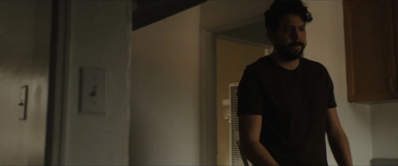 Stalker (2020) movie still: medium — A dark apartment interior features a man standing near a wall. The man is positioned in the foreground of a dim…