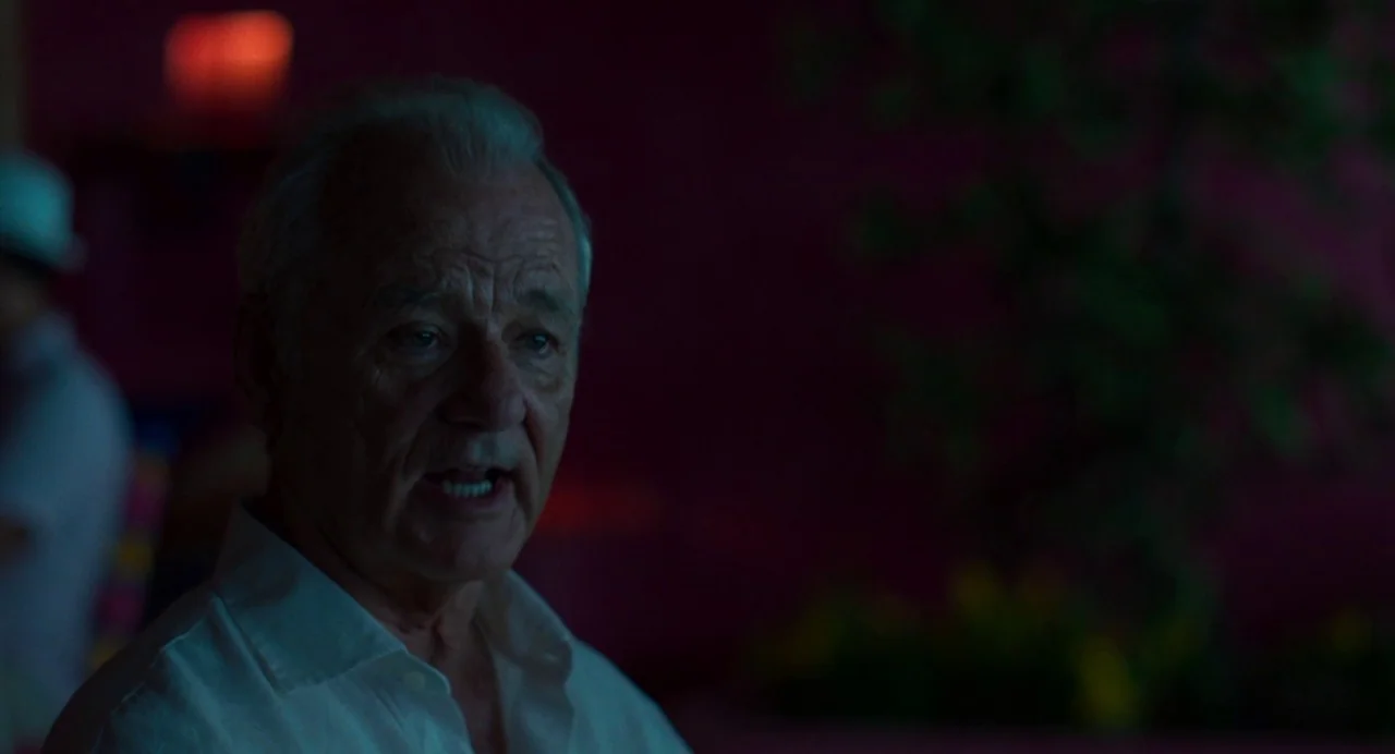 On The Rocks (2020) movie still: closeup — A dimly lit interior space with deep magenta walls and blurred foliage features. An older man in a white…
