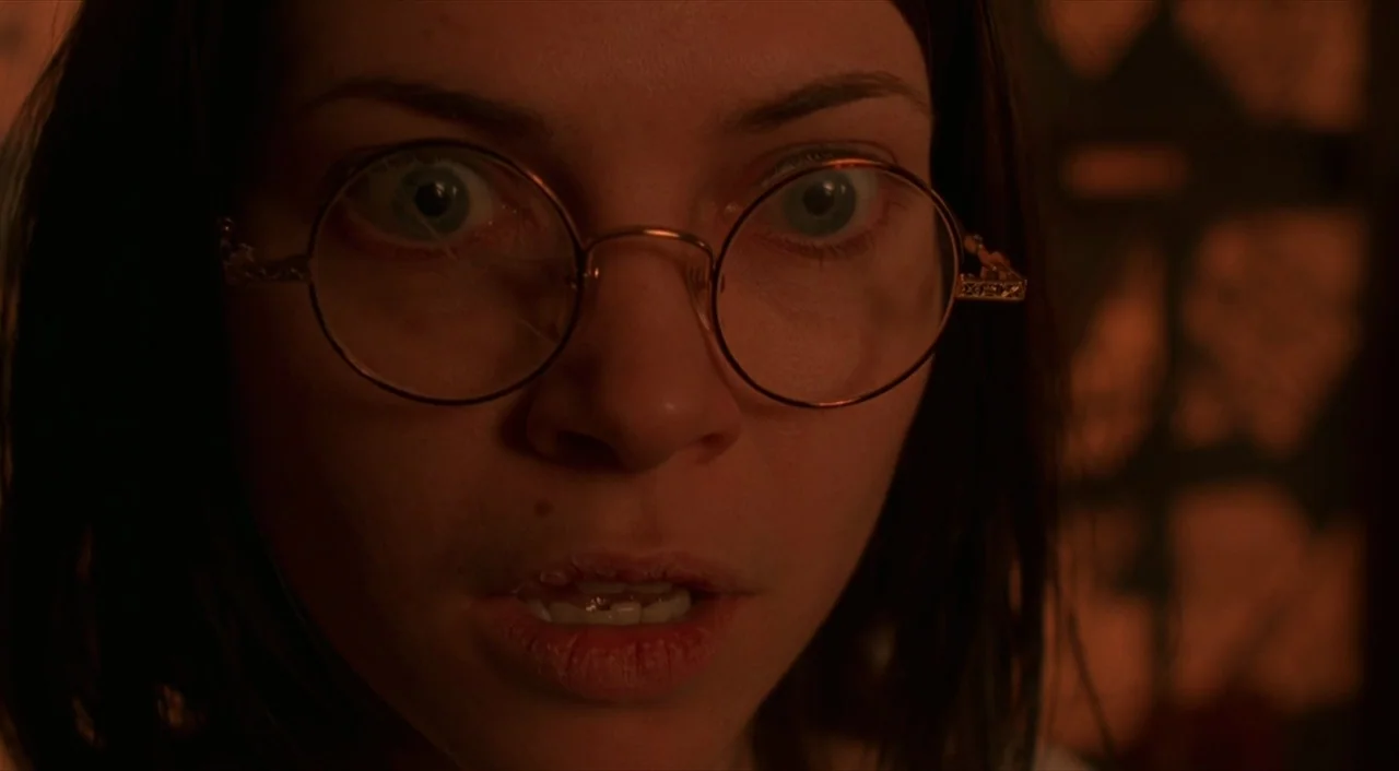 Cube (1997) movie still: closeup — A dimly lit interior space with a warm orange light hitting the subject. A young woman with dark hair wears…
