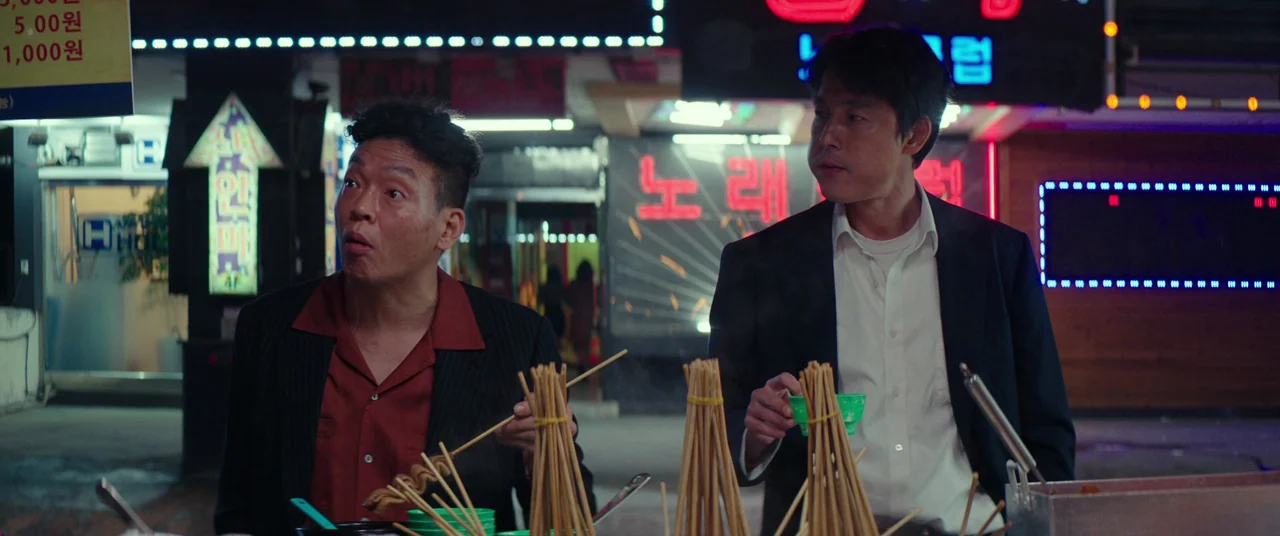 Beasts Clawing At Straws (2020) movie still: medium — A nighttime urban street features a food stall with glowing red neon signs. Two men in suits…