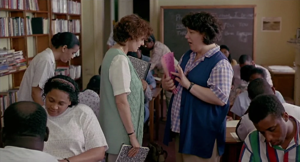Dead Man Walking (1995) movie still: medium — A crowded school classroom has wooden desks and tall bookshelves filled with books. Two middle-aged…