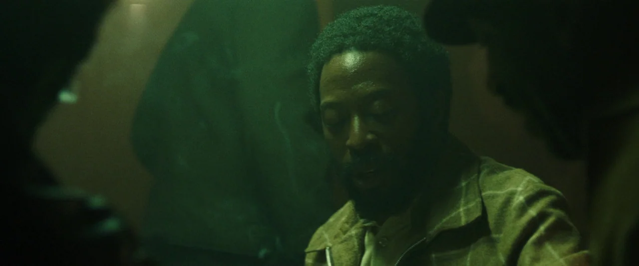 Mangrove (2020) movie still: medium — A dark space lit by a single dim green source casts deep shadows. A man with a beard stands with eyes downcast…