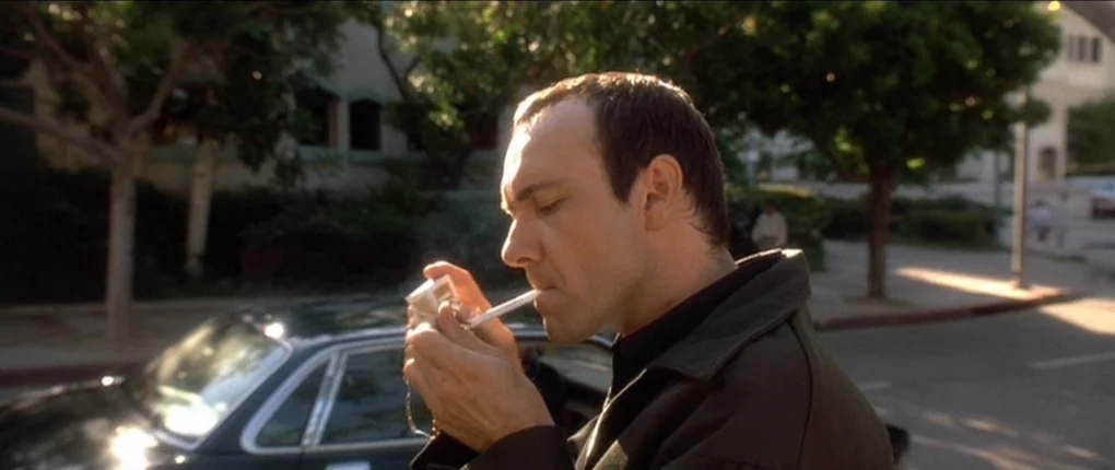 The Usual Suspects movie still: medium — A suburban street corner features mature trees and a parked black luxury sedan. A man stands in the street…
