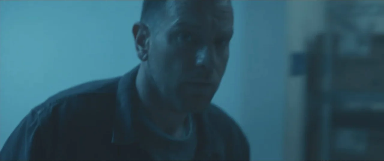 Zoe (2018) movie still: closeup — A dimly lit interior space features a man standing in blue-toned shadow. The man wears a dark shirt and gazes…