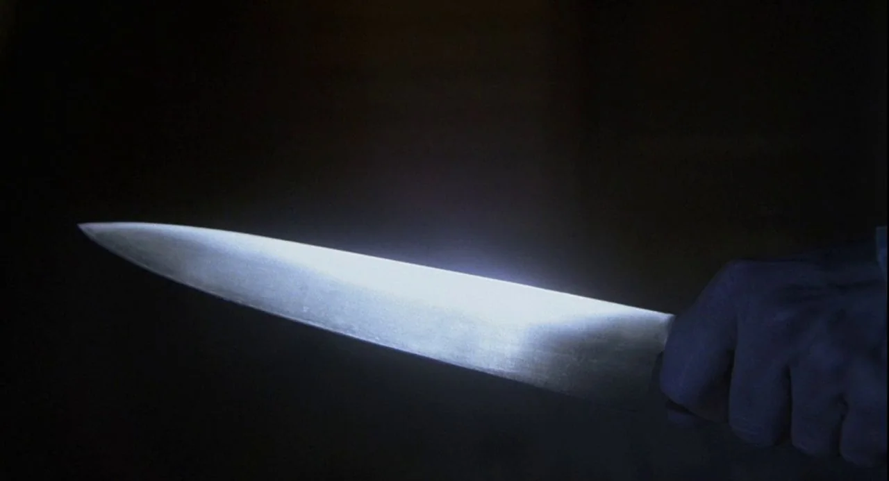 Psycho III (1986) movie still: closeup — A close up of a sharp kitchen knife held against a pitch black background. The blade is illuminated by a…
