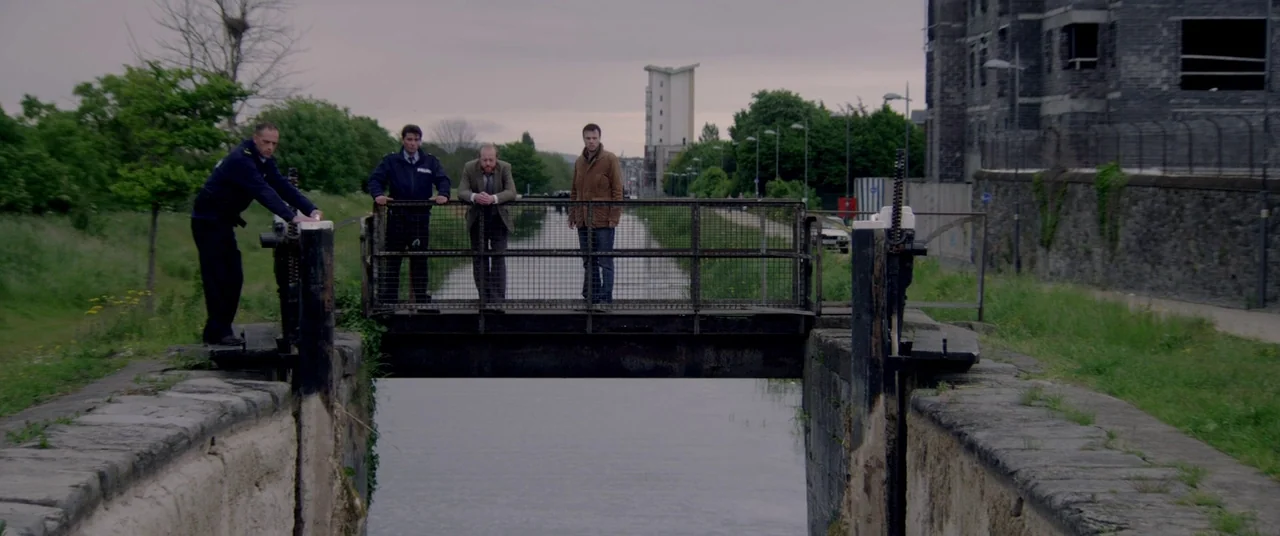 The Canal (2014) movie still: wide — A metal bridge spans a concrete canal lock surrounded by green foliage and urban structures. Four men in…
