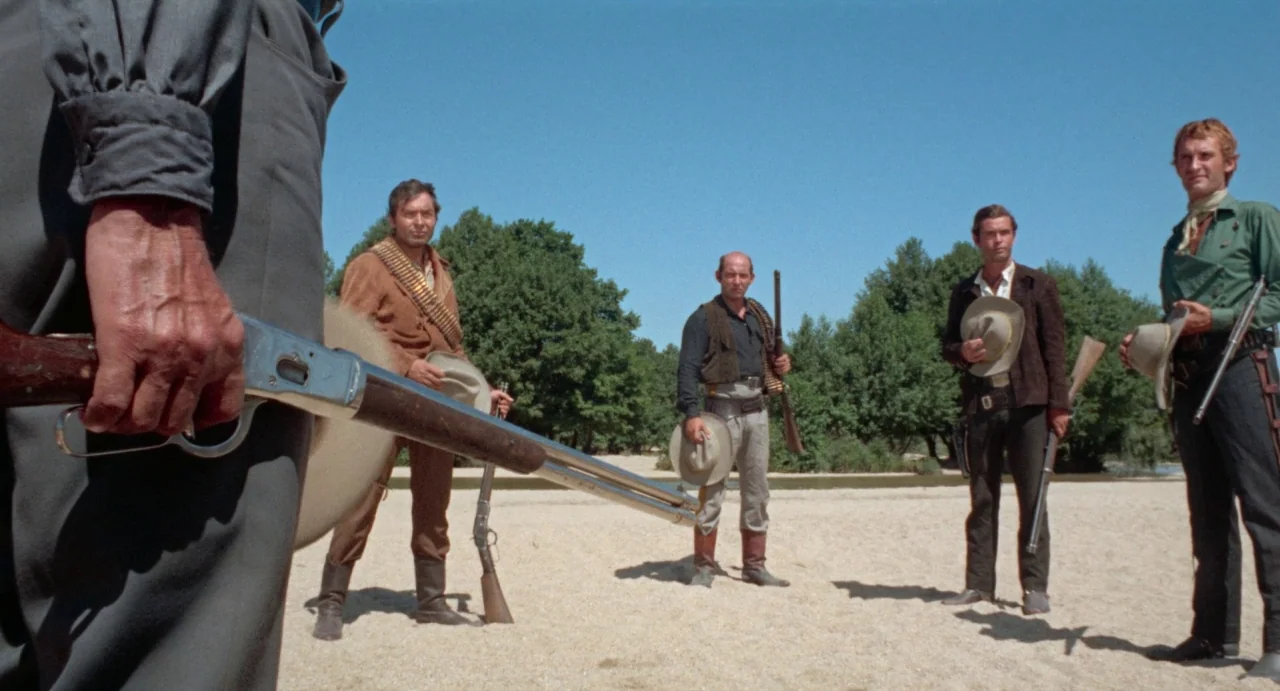 The Hellbenders (1967) movie still: fullbody — A sunny riverbank with sparse trees and open sandy ground. Four men stand in a group holding rifles…