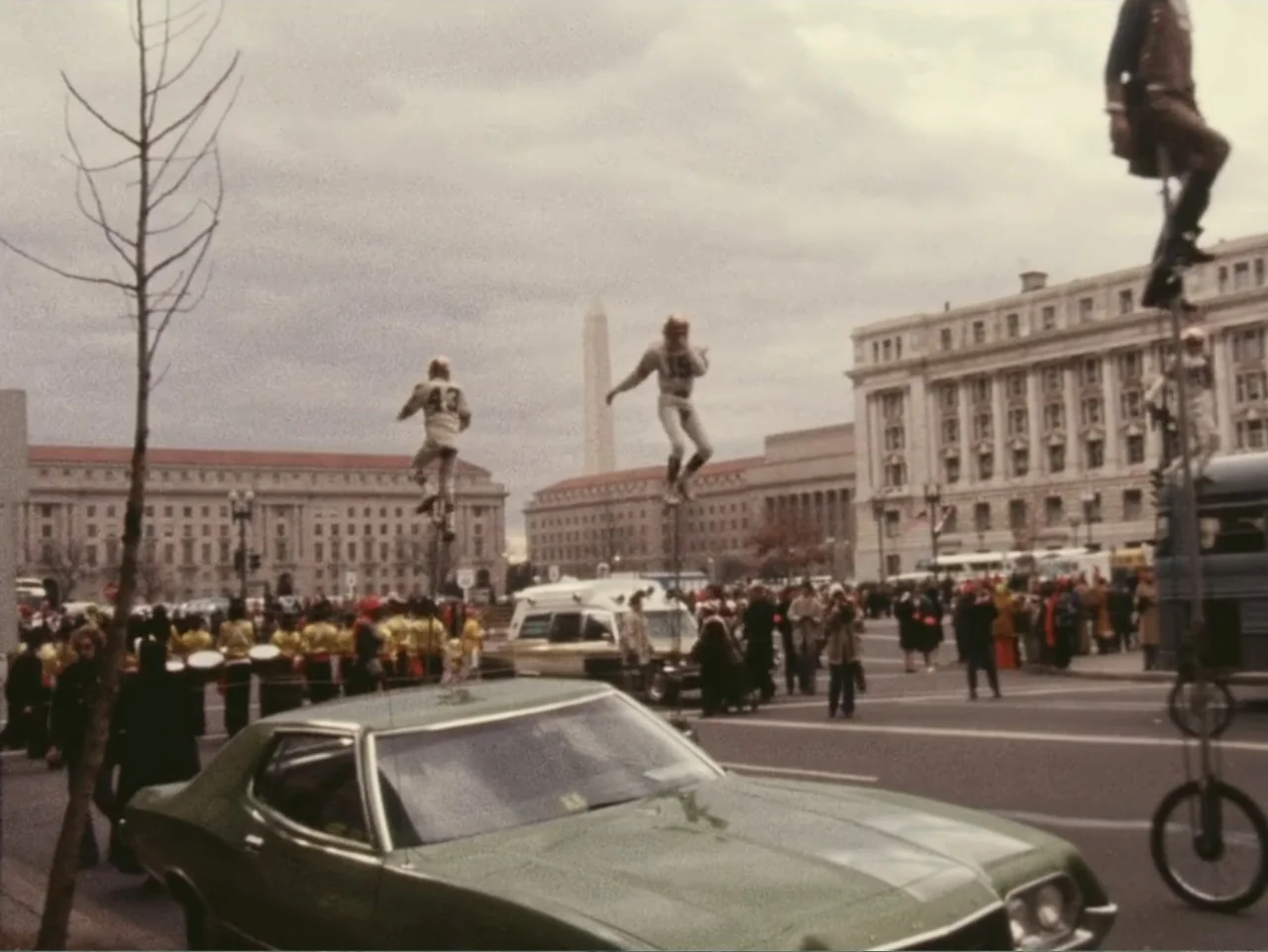 Don't Expect Too Much (2011) movie still: wide — A city street in Washington D.C. features a parade with performers on tall unicycles. A green…