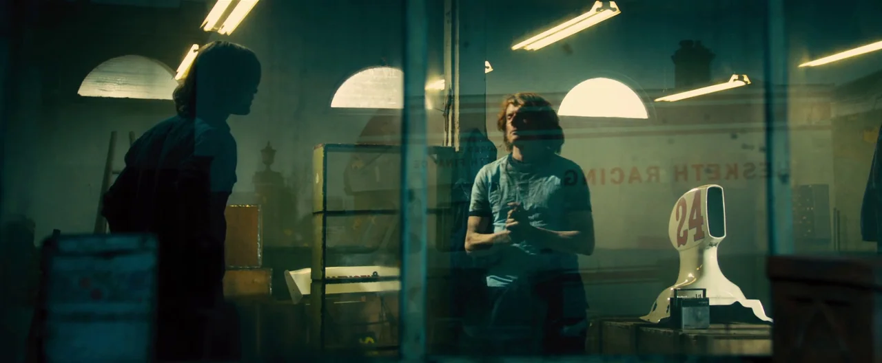 Rush (2013) movie still: medium — An industrial garage contains racing car components and clutter beneath high arched windows. Two men engage in…