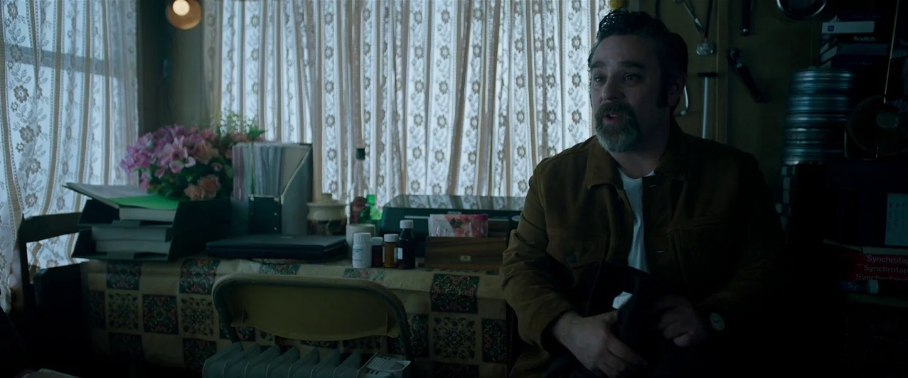 Ghost Stories (2017) movie still: medium — A cluttered room features a desk covered with office supplies in front of lace curtains. A man sits in a…