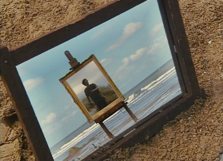 The Beaches of Agnès (2008) movie still: closeup — A beach with fine brown sand and the ocean visible in the background. An easel holds a framed…