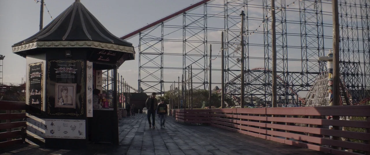 A Monster Calls (2016) movie still: wide — A wooden boardwalk at an amusement park features a small temporary hair braiding booth. An adult male and…